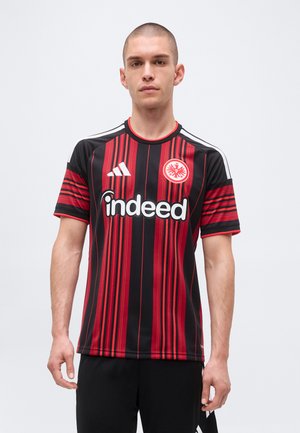 Tânăr purtând un tricou de fotbal cu dungi negre și roșii, cu logo-ul sponsorului "indeed" și emblema Eintracht Frankfurt pe piept.