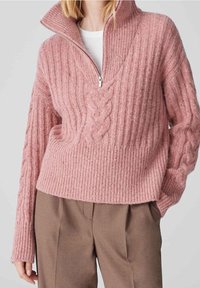 Femme portant un pull en tricot côtelé rose avec un col demi-fermeture éclair et un détail en maille torsadée, associé à un pantalon marron taille haute.