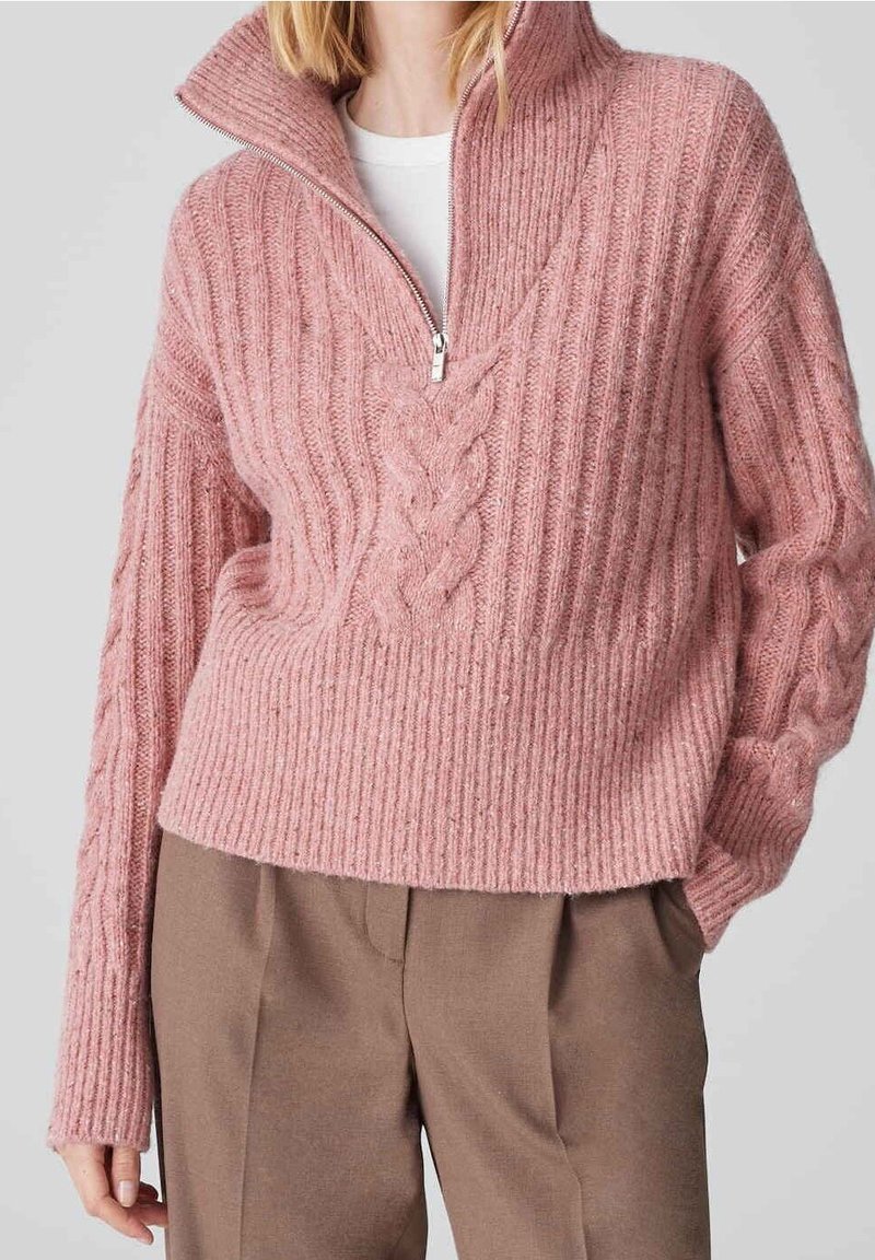 Femme portant un pull en tricot côtelé rose avec un col demi-fermeture éclair et un détail en maille torsadée, associé à un pantalon marron taille haute.