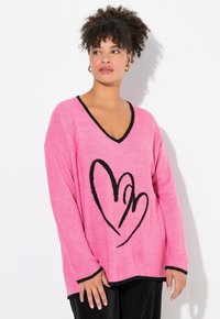 Rosa stickad tröja med svart V-ringning, prydd med en stor svart hjärtformad kontur på framsidan, och långa ärmar med svarta manschetter.
