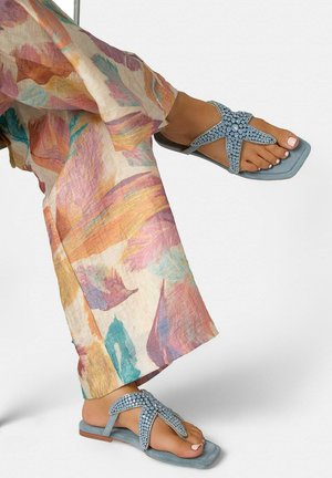 Füße, die blaue flache Sandalen mit Schmucksteinen tragen, kombiniert mit bunten, locker sitzenden Hosen mit abstrakten Pastellmustern.
