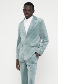 Fato de veludo azul claro com dois botões, lapelas recortadas e bolsos laterais. Modelo posa com as mãos nos bolsos contra um fundo branco.