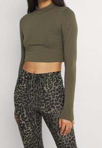 Olivegrünes, kurzes Langarmoberteil, kombiniert mit enganliegenden Leggings mit einem dunklen Leopardenmuster. Der Stoff wirkt glatt und dehnbar.