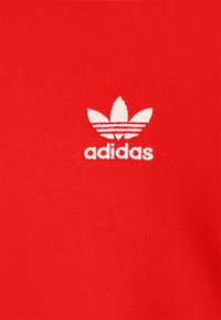 Logo trefoil Adidas blanc brodé sur un fond en tissu texturé rouge vif.