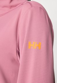 Μωβ μπλούζα με μακριά μανίκια και υφή υφάσματος· διαθέτει πορτοκαλί λογότυπο "HH" στο αριστερό μανίκι. Ορατές ραφές λεπτομέρειες.