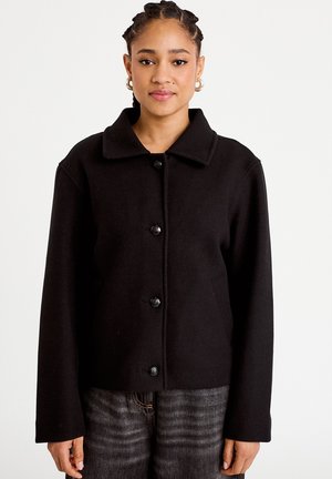 Kvinde med fletninger iført guldringe og en sort jakke med knapper over grå jeans mod en hvid baggrund.