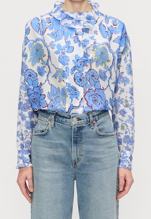 Kobieta ubrana w niebiesko-białą kwiecistą bluzkę z długim rękawem z falbaną przy kołnierzu i mankietach, zestawioną z wysokim stanem jasnoniebieskich dżinsów.