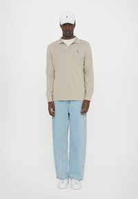 Polo de manga longa bege claro, boné branco, jeans largos azul claro e ténis brancos. Apresenta um pequeno logo no polo.