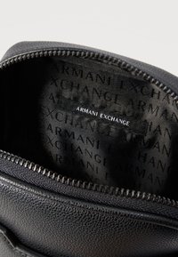 Μαύρη τσάντα από υφή δέρματος, μερικώς ανοιγμένη, που αποκαλύπτει το εσωτερικό ύφασμα με τύπωμα «Armani Exchange» και μια μαύρη ετικέτα με λευκό κείμενο «Armani Exchange».