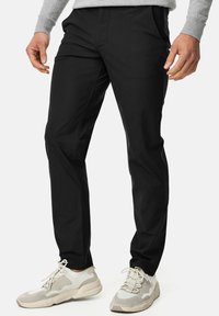Schwarze Hosen mit glatter Textur, ausgestattet mit Fronttaschen und schmalem Schnitt, kombiniert mit hellen Sneakers für einen lässigen Look.