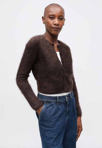 Cardigan marrón peludo con cuello redondo, mangas largas y botones frontales, combinado con vaqueros de mezclilla azul de cintura alta.