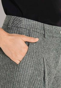 Hand rustend in de zak van grijs gestreepte broek, gecombineerd met een zwarte top, waarbij de stofstructuur en de details van de zak zichtbaar zijn.