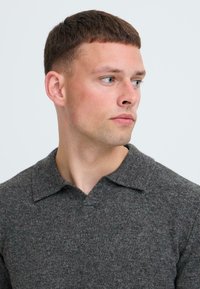 Grauer Pullover mit Polokragen, strukturiertem Stoff, sichtbaren Nähten und einem kantigen Halsausschnitt. Das Modell hat kurze Haare und schaut zur Seite.