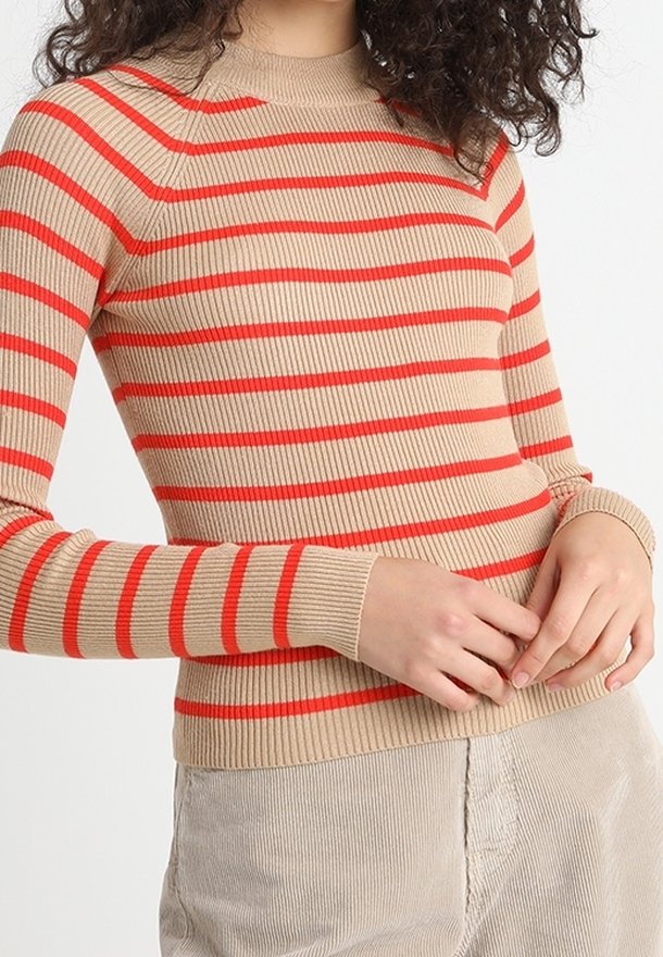 Haut beige côtelé à manches longues avec des rayures rouges horizontales porté par une personne aux cheveux bouclés et foncés, associé à un pantalon beige clair.