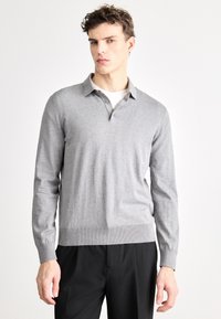 Tricou polo gri cu mâneci lungi și guler, având o textură netedă și tiv striat. Purtat peste un tricou alb și pantaloni negri.