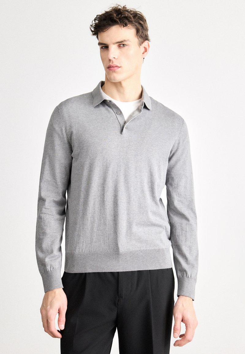 Tricou polo gri cu mâneci lungi și guler, având o textură netedă și tiv striat. Purtat peste un tricou alb și pantaloni negri.