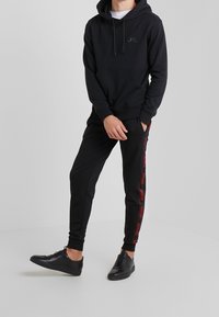 Schwarzer Hoodie mit Kängurutasche und kleinem Logo. Schwarze Jogginghose mit rotem Seitenstreifen. Schwarze Sneakers.