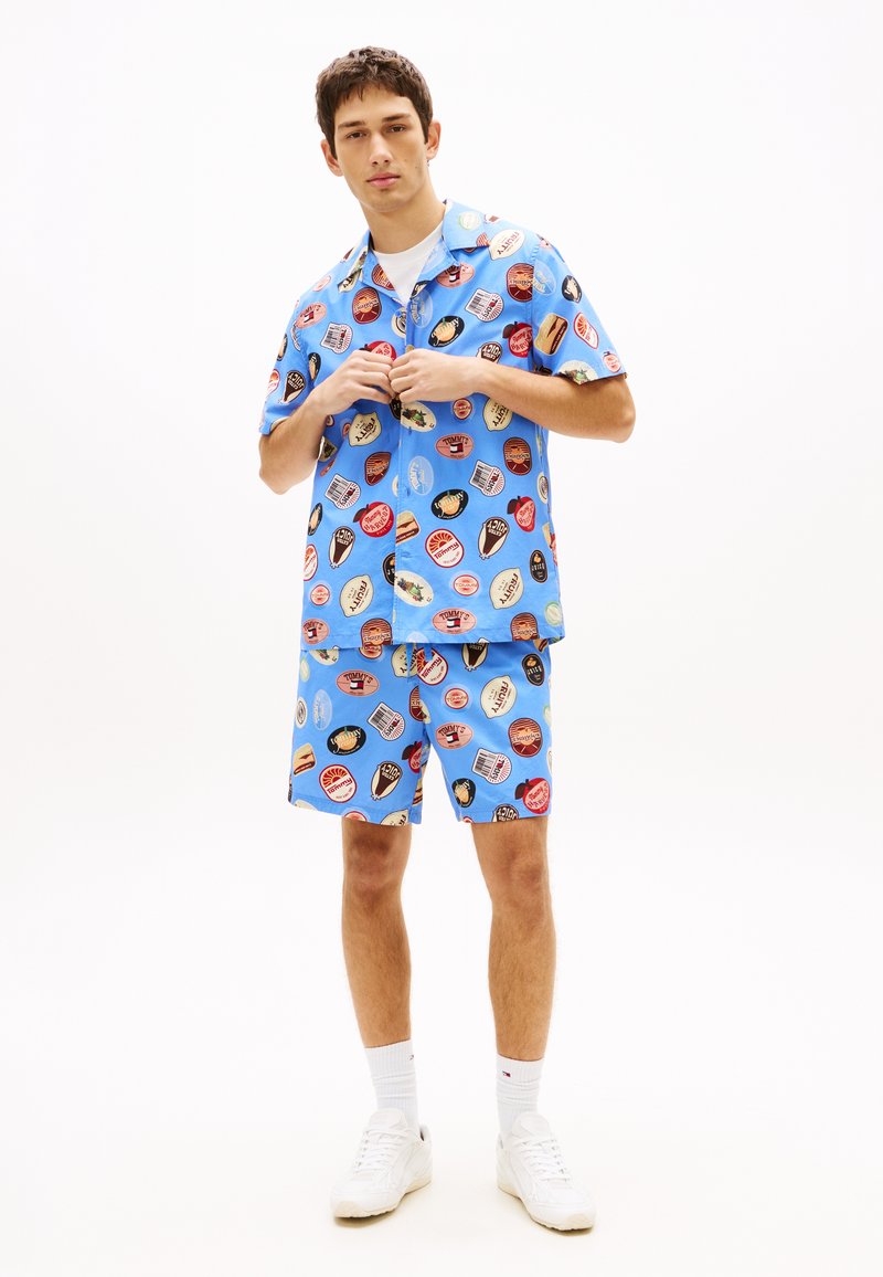 Jeune homme debout portant une chemise à manches courtes bleue assortie à un short, tous deux ornés d'imprimés graphiques circulaires colorés, des baskets blanches et des chaussettes.
