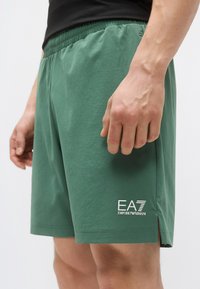Žalios medvilninės šortų su elastinga juosmens juosta, šoninėmis skeltukėmis ir baltu logotipo detalės užrašu „EA7 EMPORIO ARMANI“ ant apatinės kojos dalies.