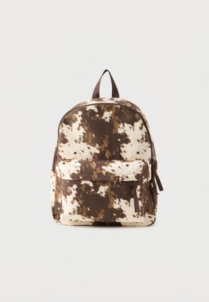 Sac à dos avec motif en peau de vache marron et crème, poche avant zippée, poignée supérieure et bretelles ajustables sur un fond uni.