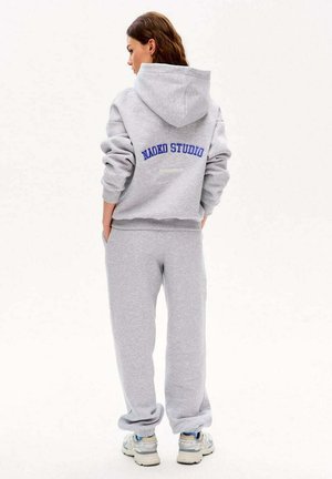 Kobieta stojąca tyłem, ubrana w jasnoszary kaptur i spodnie dresowe z napisem "NAOKO STUDIO" w niebieskim kolorze na plecach kaptura.