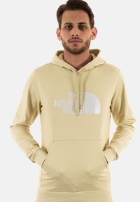 Sudadera con capucha beige con un bolsillo tipo canguro en el frente, que presenta un gran estampado blanco del logotipo "The North Face" en el pecho.