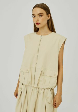 Beige mouwloze vest met een rits aan de voorkant, voorzien van twee voorzakken en een gegatherde zoom. Gemaakt van een gladde stof met een effen textuur.