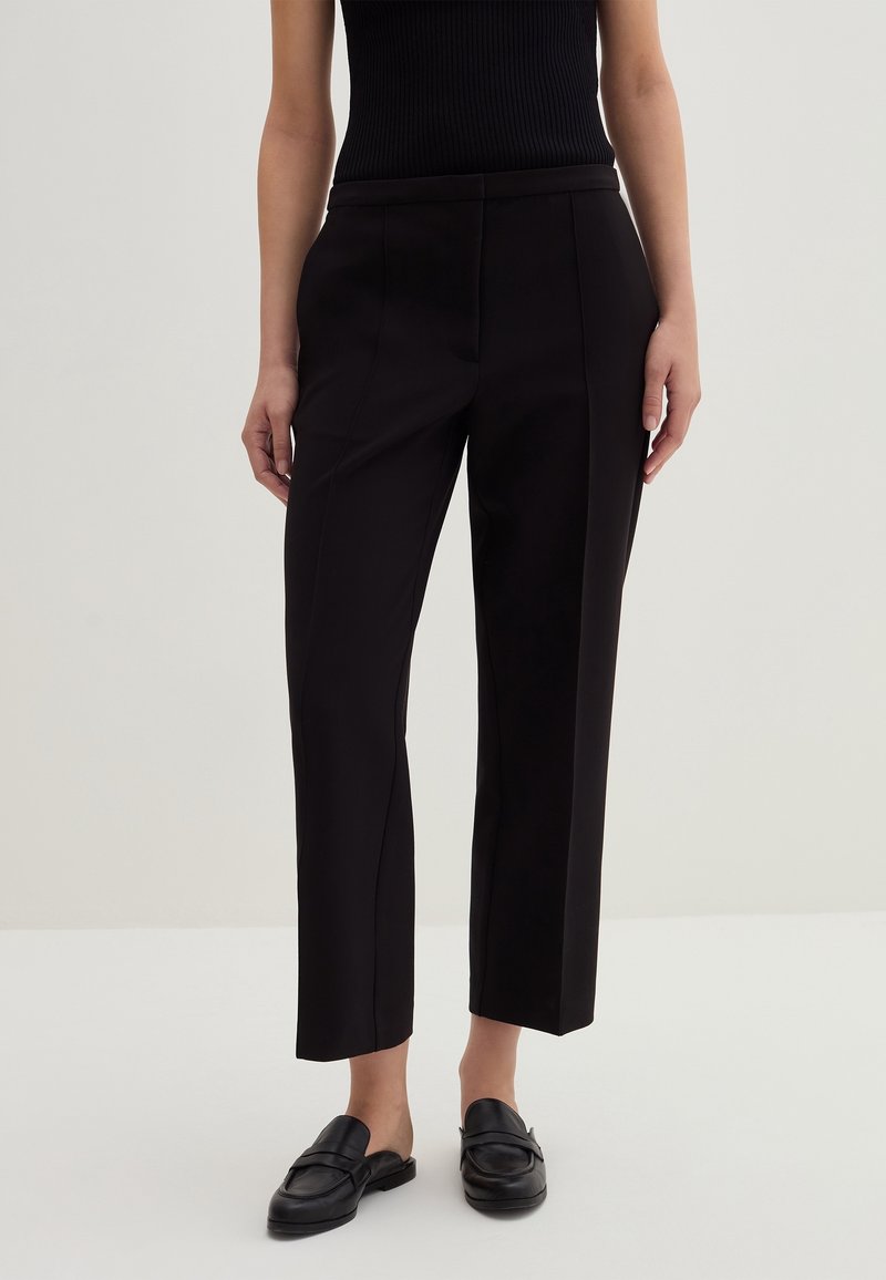 Femme portant un pantalon court noir, un haut noir sans manches et des mocassins noirs sans lacets, debout devant un fond uni.