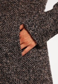 Textureret, flerfarvet frakke i mørkegrå og brun med en hyggelig fornemmelse. Har en sidelomme og en synlig hånd, der holder i lomme kanten.