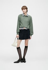 Joven vistiendo un suéter verde sobre una camisa con cuello, falda negra con pliegues, medias transparentes hasta la rodilla, tacones negros, sosteniendo un bolso blanco.