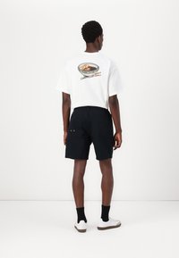 Hvid t-shirt med en skål grafik og pinde-design på ryggen, parret med sorte shorts og hvide sneakers.