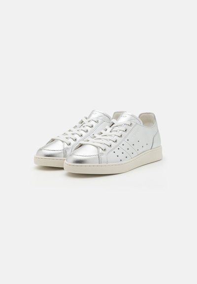 Silver vegan leather sneakers with circular perforations, rounded toe, and neutral rubber sole. Laces and branding detail on tongue.
