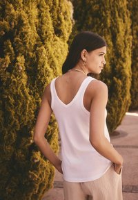 Hvid, ærmeløs tanktop med en V-formet ryg, lavet af blødt stof. Har et enkelt design, iført med lyse, strukturerede bukser.