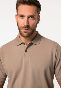 Beige polo tričko vyrobené z texturované látky, má klasický límec, zapínání na tři knoflíky a krátké rukávy s volným střihem.