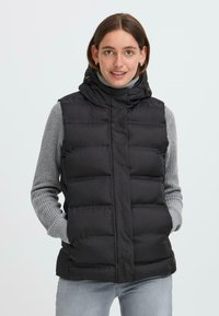 Une jeune femme sourit, portant un gilet en duvet noir par-dessus un col roulé gris, avec les mains dans les poches et un fond neutre.