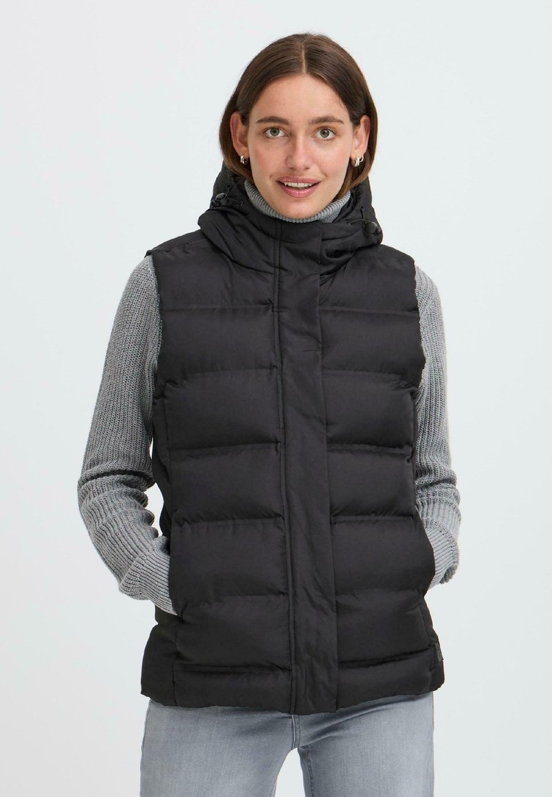 Une jeune femme sourit, portant un gilet en duvet noir par-dessus un col roulé gris, avec les mains dans les poches et un fond neutre.