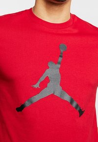 Camiseta de algodón roja con una gran silueta negra de una figura saltando que sostiene un balón de baloncesto, con cuello acanalado y mangas cortas.