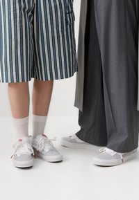 Weiße und graue Turnschuhe mit roten Akzenten verfügen über ein Obermaterial aus Mesh und Synthetik, eine abgerundete Spitze und flache Schnürsenkel, kombiniert mit gestreiften und grauen Hosen.