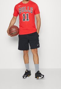 Červené tričko s krátkým rukávem s nápisem "BULLS" a číslem "11" v černé barvě. Černé šortky, šedé ponožky a černé sportovní boty se zlatými akcenty, držící basketbal.