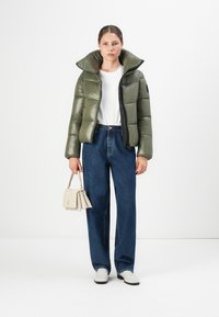 Olijfgroene pufferjack met een hoge kraag, ritssluiting en zwarte accenten, gecombineerd met brede blauwe spijkerbroeken en witte schoenen.