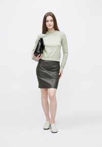 Svetlo zelena pletena jopica z okroglim izrezom, kombinirana s temno usnjeno svinčnikasto krilo. Model drži črno torbico in nosi svetle loaferje.