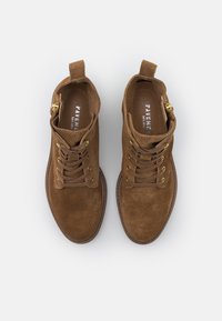 Bottines en daim marron avec un bout arrondi, des œillets dorés et des lacets. Elles sont dotées d'une boucle de tirage et d'une semelle intérieure texturée pour le confort.