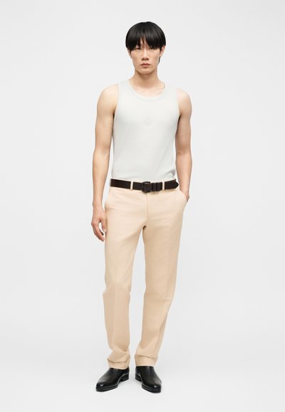 Νεαρός άνδρας όρθιος, φορώντας λευκό αμάνικο πουκάμισο, μπεζ παντελόνι με μαύρη ζώνη και μαύρα παπούτσια, σε απλό φωτεινό φόντο.