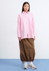Mladá žena stojí ve studiu, oblečená v růžovo-bílém pruhovaném oversized triku a hnědé sukni, které doplňuje stříbrnými teniskami.