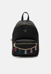 Čierny prešívaný batoh so zlatým kovovaním, ktorý má predný zipsový vrecko ozdobené viacfarebnými písmenami, ktoré hlásia "GUESS" na retiazke.
