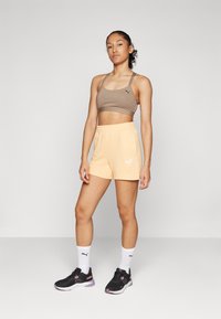Lätta persikofärgade shorts och en brun crop top, båda med Puma-logotypen. Modellen har på sig svarta sneakers med rosa detaljer och vita strumpor.