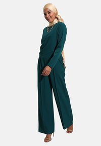 Mono azul verdoso con mangas largas y un diseño de cuerpo drapeado. Presenta piernas anchas, textura suave y un collar de cadena dorada como acento.