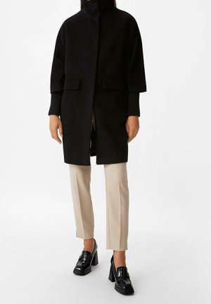 Kobieta w czarnym płaszczu o długości do połowy uda, beżowych spodniach do kostek i czarnych mokasynach na szerokim obcasie, stojąca na białym tle.