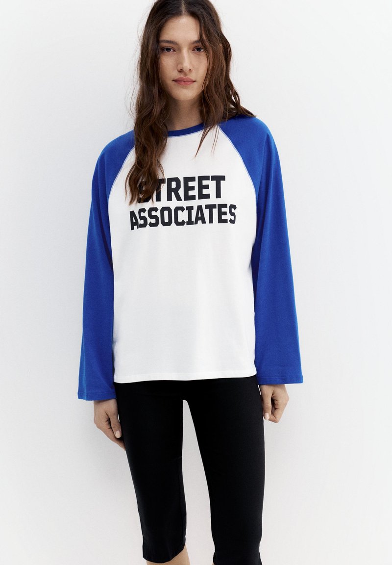 Jeune femme aux longs cheveux bruns portant un t-shirt à manches longues blanc et bleu avec le texte "STREET ASSOCIATES" et un pantalon noir, debout contre un mur blanc.