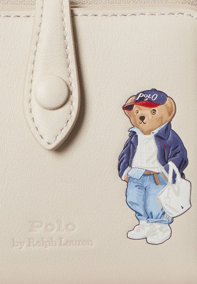 Κρεμ δερμάτινο υλικό με πολύχρωμη εικονογράφηση μιας αρκούδας που φορά μπλε μπουφάν και καπέλο, κρατώντας μια λευκή τσάντα. Χαραγμένο το "Polo by Ralph Lauren".
